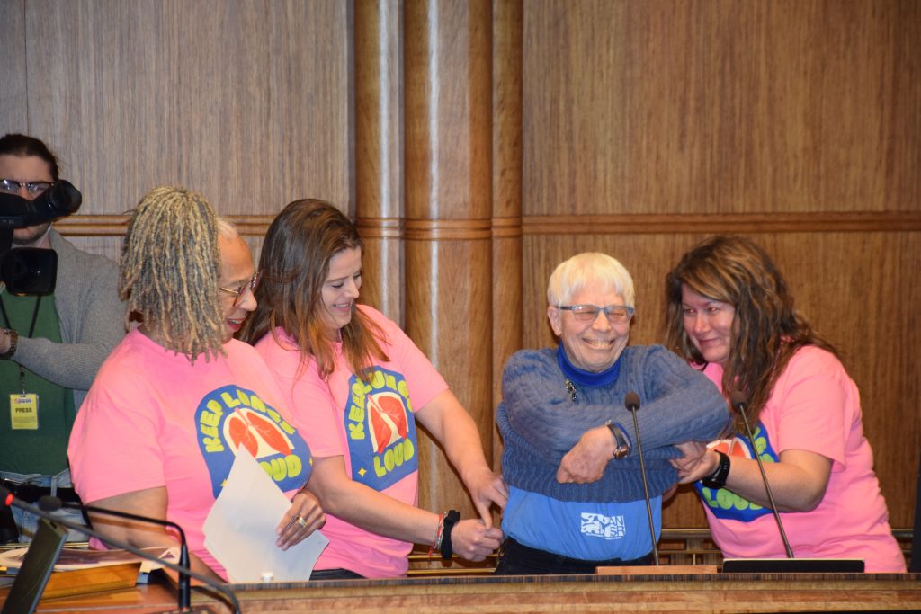 Three women in pink shirts help another woman in blue remove her sweater. They stand at a podium. A cameraman stands behind them.