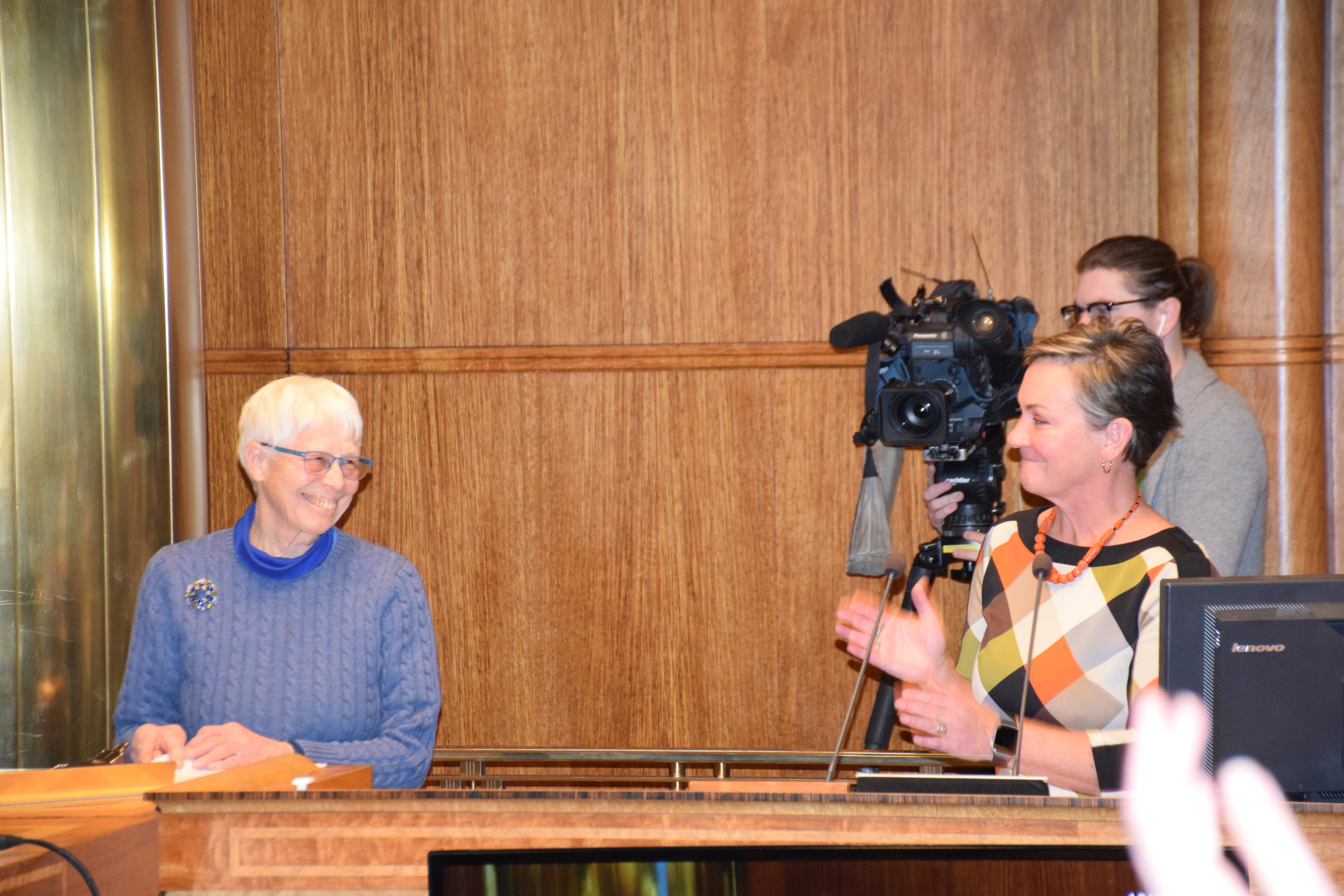 A woman applauds another woman to her right while standing at a podium. The woman on the right smiles back. Behind them, a cameraman.